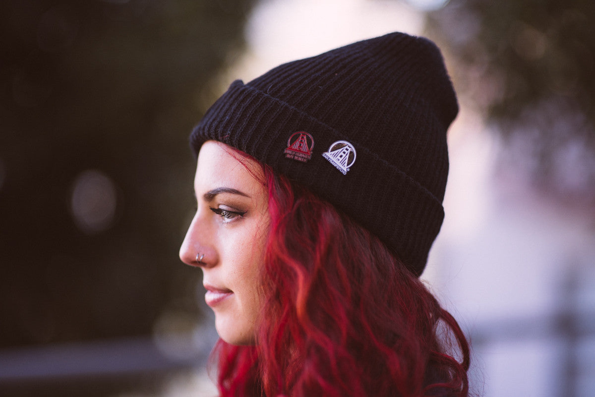 A person with red hair, wearing a black beanie and a nose piercing, faces sideways outdoors. Adorning their jacket is the Black & White SFP Logo Pin that glistens among a striking collection of custom enamel pins, each piece narrating its own vibrant story.