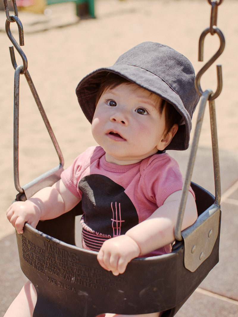 A baby dressed in a Sutro Tower Onesie, adorned with a pastel pink color and matching hat, sits curiously in a swing at the playground. This playful outfit is crafted from 100% cotton, ensuring comfort during their little adventure.