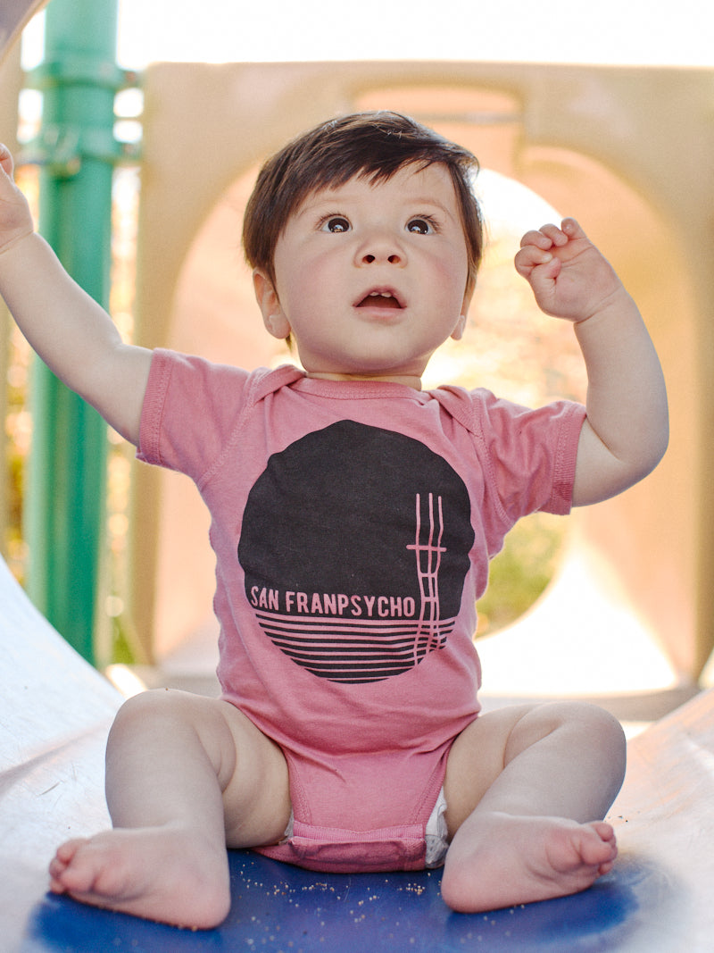 A baby in a pastel pink Sutro Tower Onesie, adorned with a fun San Franchpsycho design, sits on a slide, gazing upwards and raising their arms.