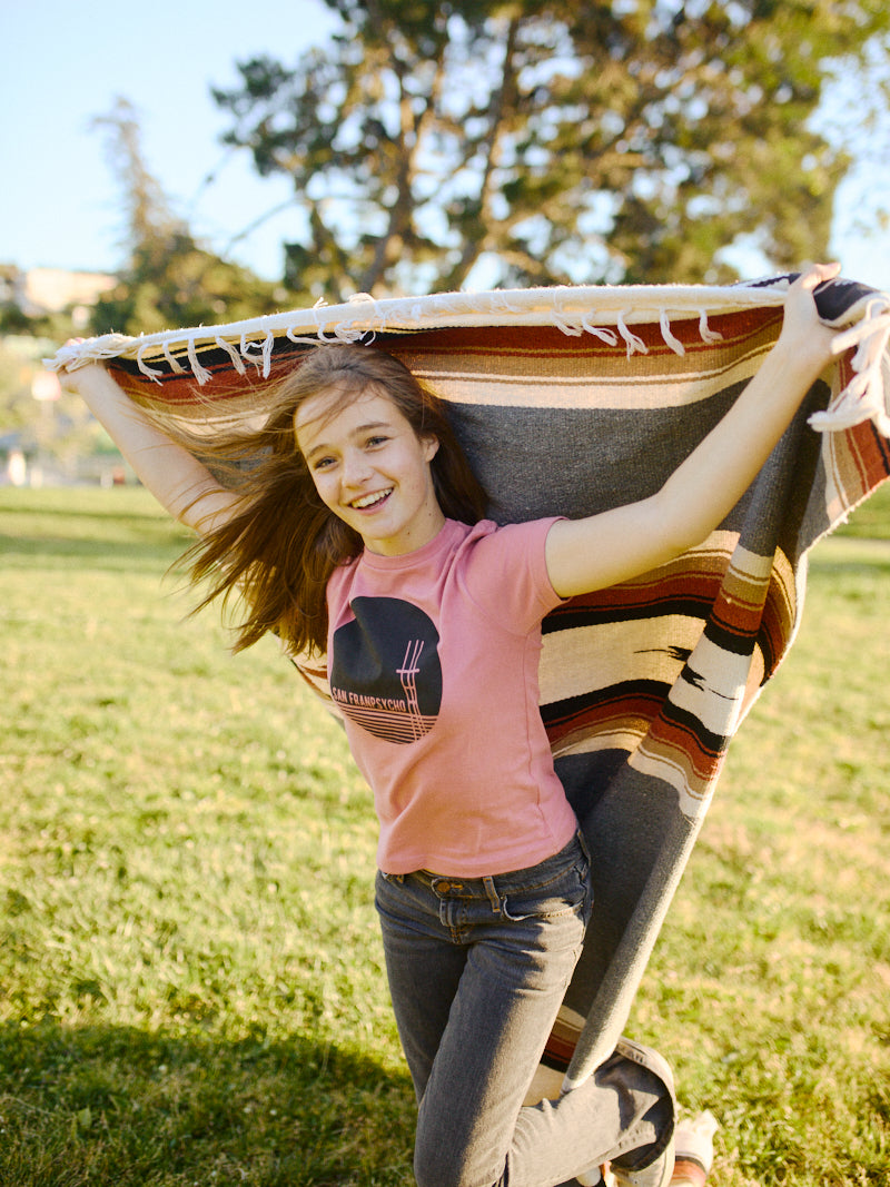 A person joyfully running with a striped blanket and wearing a Youth Sutro Tower Tee in pastel pink, soaking up the sun in a park on a perfect day.
