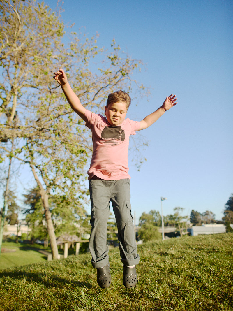 A child jumps joyfully in the park, wearing a pastel pink Youth Sutro Tower Tee and gray pants, with trees and a clear blue sky in the background.