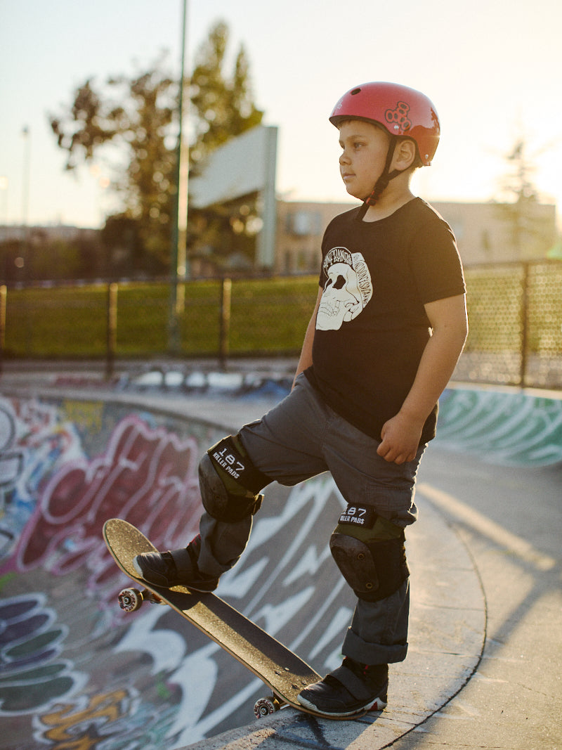 A child in a Youth Black Street Skull Tee and skate gear stands on a skateboard at the skatepark, wearing a helmet and kneepads.
