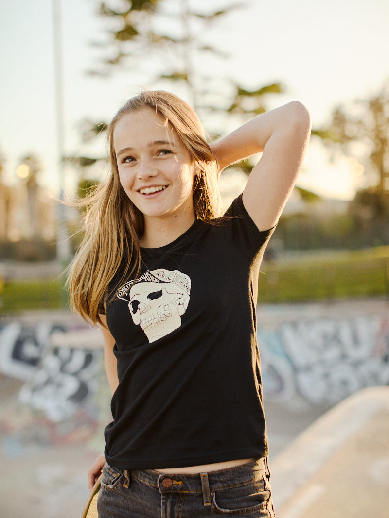 A smiling girl in the Youth Black Street Skull Tee stands outside, framed by trees and vibrant graffiti.