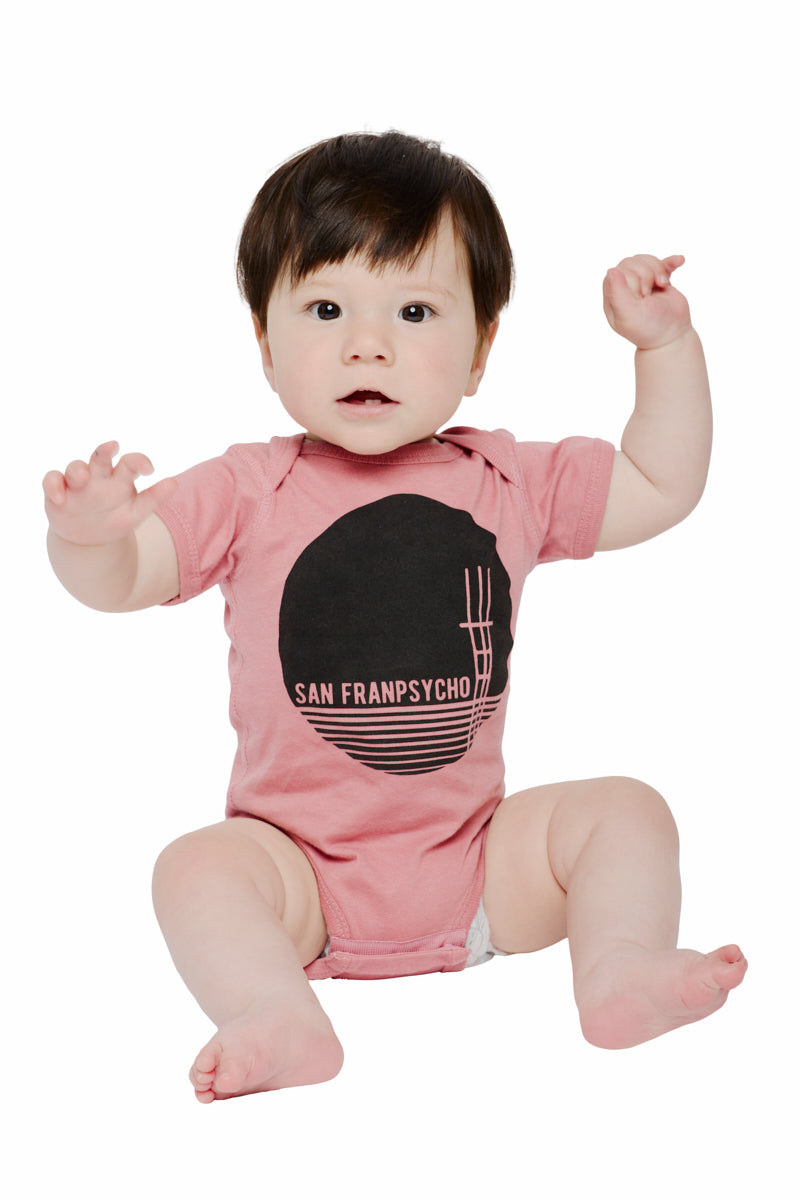 A baby in a pastel pink Sutro Tower Onesie featuring the San Franpsycho logo sits with raised arms against a white background. The 100% cotton fabric guarantees comfort, while a subtle print of the Sutro Tower adds an iconic touch of charm.