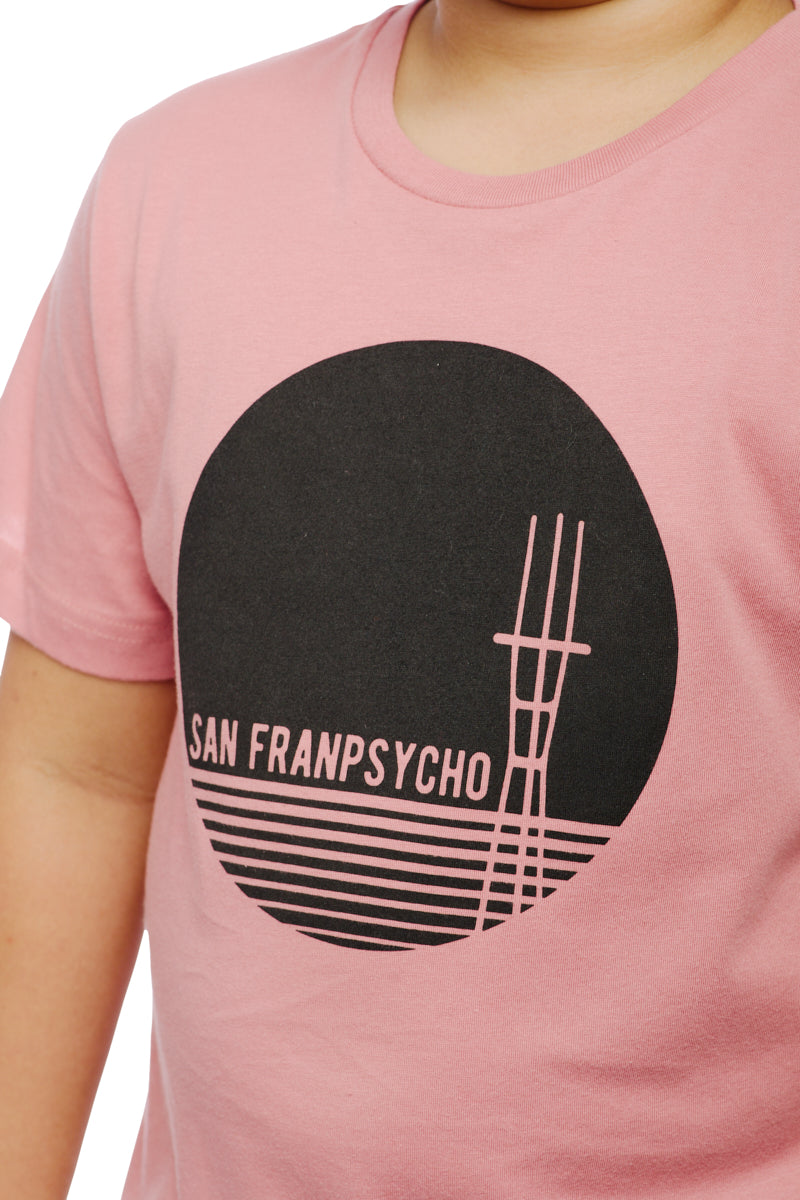 A child wears a pastel pink Youth Sutro Tower Tee, showcasing a black circle design and the iconic San Franpsycho text, all made from 100% cotton.