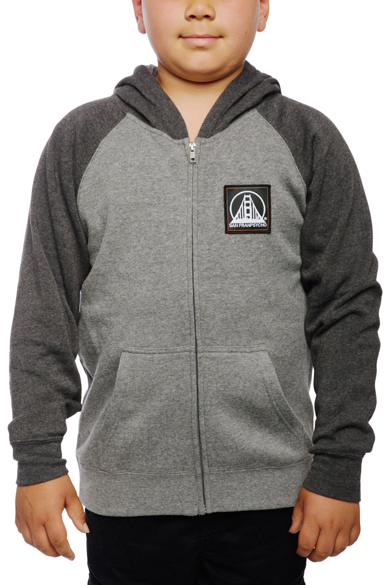 A child wearing the Youth Two Tone Patch Hoodie, featuring a bridge logo on the chest, stands against a white background, showcasing the perfect blend of style and comfort found in San Franpsycho's gray youth hoodies.