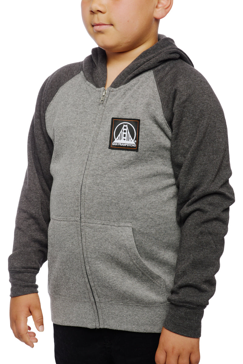A child wears the Youth Two Tone Patch Hoodie, featuring a gray and black design with a bridge logo on the chest, embodying iconic San Franpsycho style. It's ideal for those looking for trendy youth hoodies that seamlessly combine urban flair with comfort.