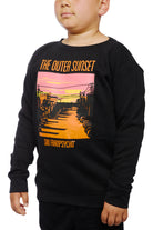 A child wears the Youth Outer Sunset Crewneck, a black sweatshirt adorned with a colorful sunset graphic labeled The Outer Sunset, San Franpsycho. This distinctive piece is hand-printed in San Francisco, highlighting local artistry and the vibrant spirit of the city.