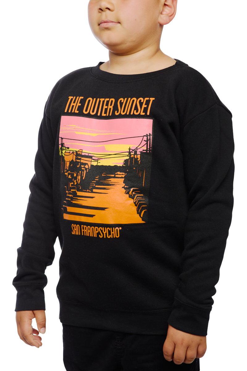 A child wears the Youth Outer Sunset Crewneck, a black sweatshirt adorned with a colorful sunset graphic labeled The Outer Sunset, San Franpsycho. This distinctive piece is hand-printed in San Francisco, highlighting local artistry and the vibrant spirit of the city.