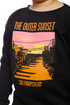 A child wears the Youth Outer Sunset Crewneck, adorned with a vibrant sunset design and "The Outer Sunset San Franpsycho" text, meticulously hand printed to reflect the enchanting views of San Francisco.