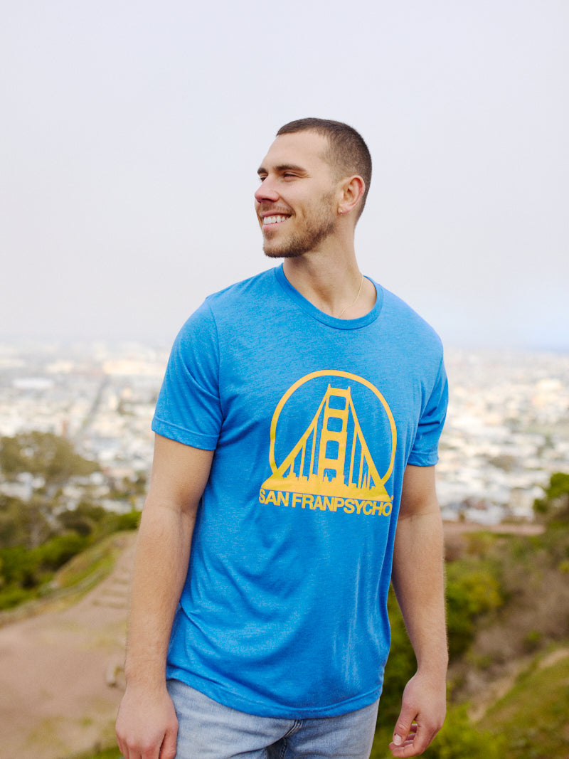 A man in a Blue & Gold Logo Tee stands outdoors, smiling against a blurred cityscape backdrop.