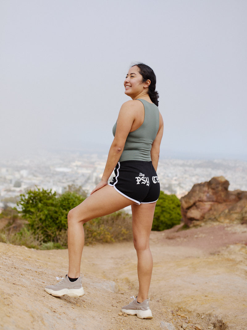 A woman in Stay Psycho Booty Shorts - Black smiles in athletic wear, standing on a rocky trail and enjoying the cozy, lightweight feel with a cityscape behind her.