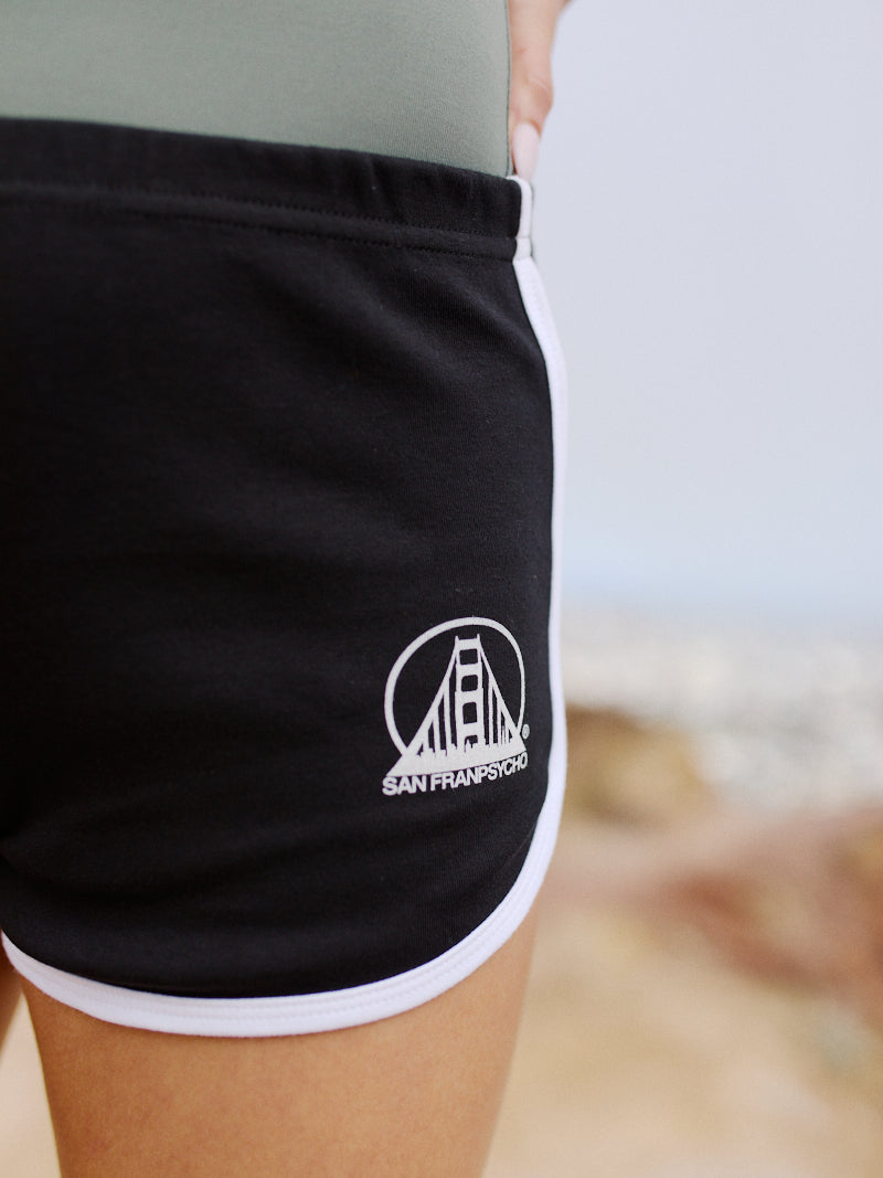 Close-up of cozy, lightweight Stay Psycho Booty Shorts - Black, featuring a white San Francisco bridge logo and white trim, worn outdoors.
