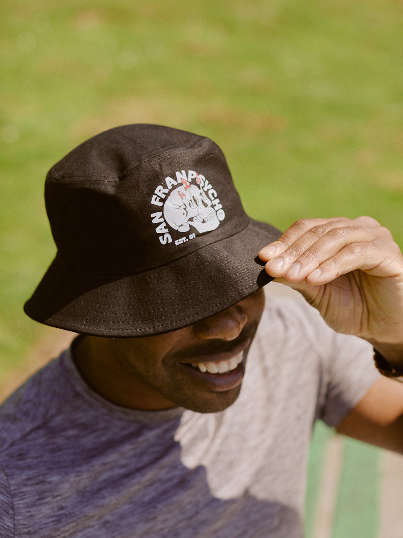 A person smiling in the outdoors, wearing a hand-printed black Shroom Skull Bucket Hat made of soft cotton, capturing the essence of San Francisco's unique charm.