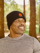Smiling man in a gray shirt and Black Lumberjack Beanie w/ Sutro Logo standing outdoors among tall trees.