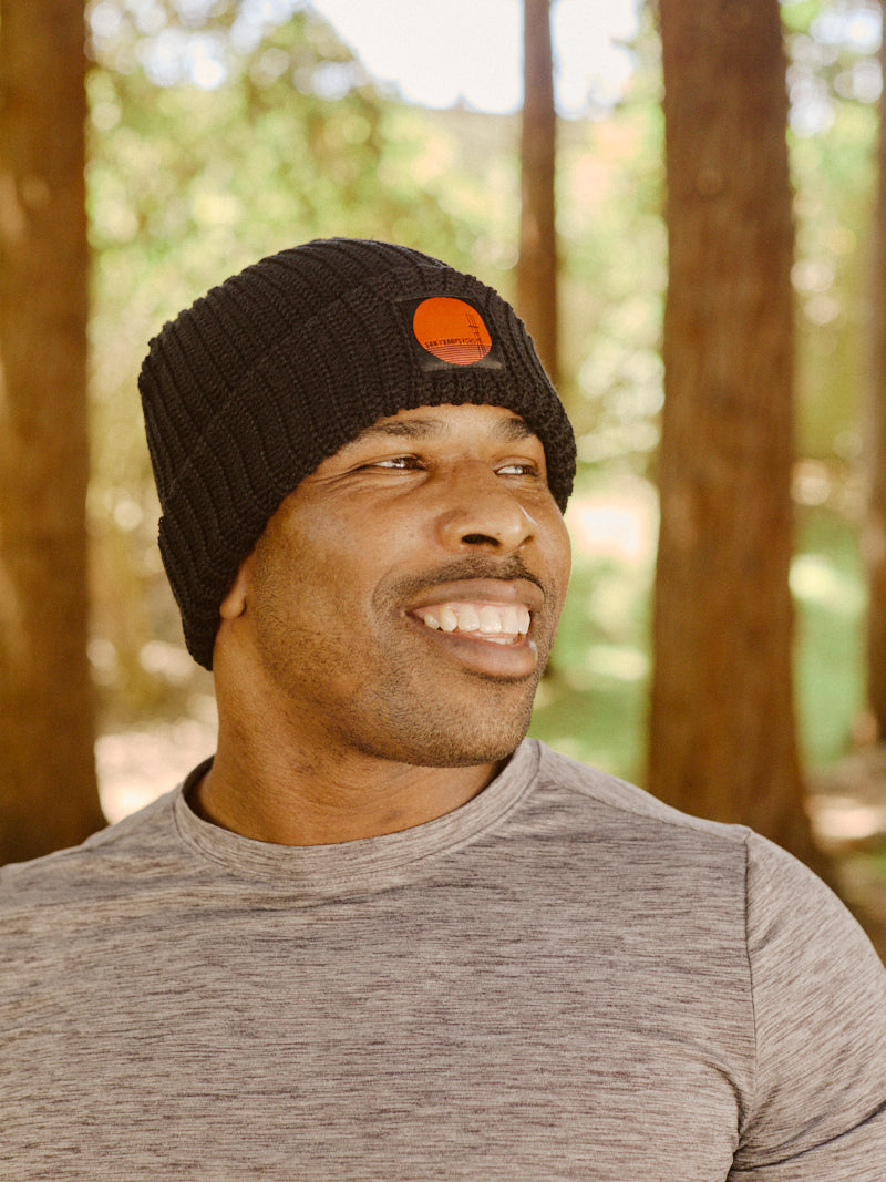 Smiling man in a gray shirt and Black Lumberjack Beanie w/ Sutro Logo standing outdoors among tall trees.