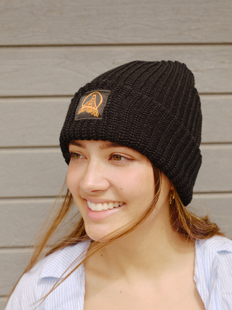 A smiling person wearing a Black Lumberjack Beanie with an Acid Logo stands in front of a wooden wall.
