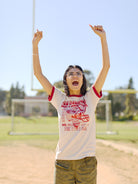 A person joyfully raises their arms on a sunny day at a sports field, wearing the vintage-inspired Candlestick Champs Ringer Tee and glasses.
