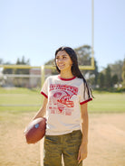 A person stands on a sunny field, proudly holding a football and sporting the Candlestick Champs Ringer Tee, ready for the Niners season.