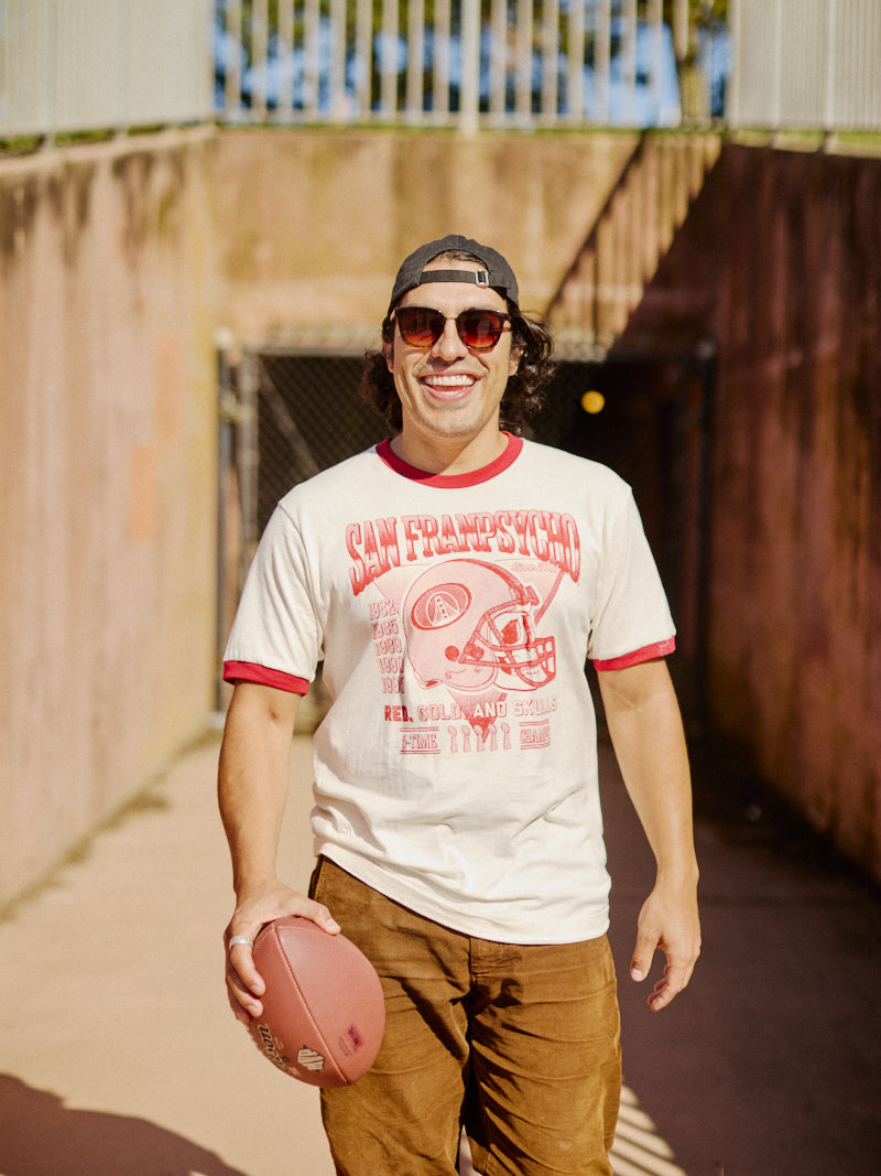 A person in a Candlestick Champs Ringer Tee and sunglasses, holding a football, stands outside in the sunlight, ready for Niners season.