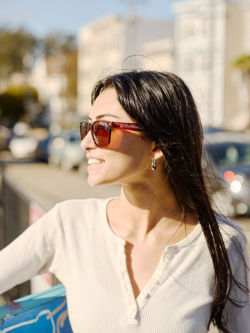 A person with long dark hair is wearing SFP Sunglasses and a white shirt, smiling outdoors on a sunny day.