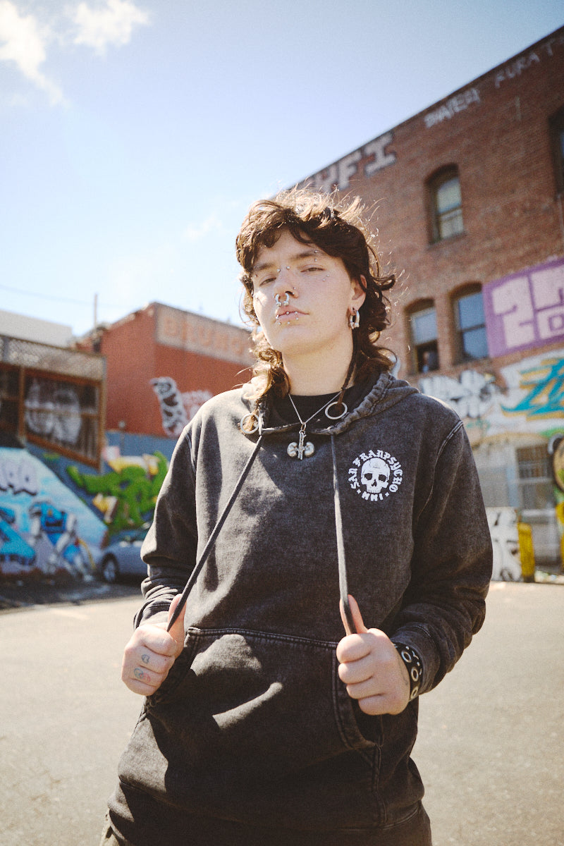 A person with piercings and curly hair is wearing a BBNW Skull Pullover Hoodie, standing on an urban street covered in graffiti.