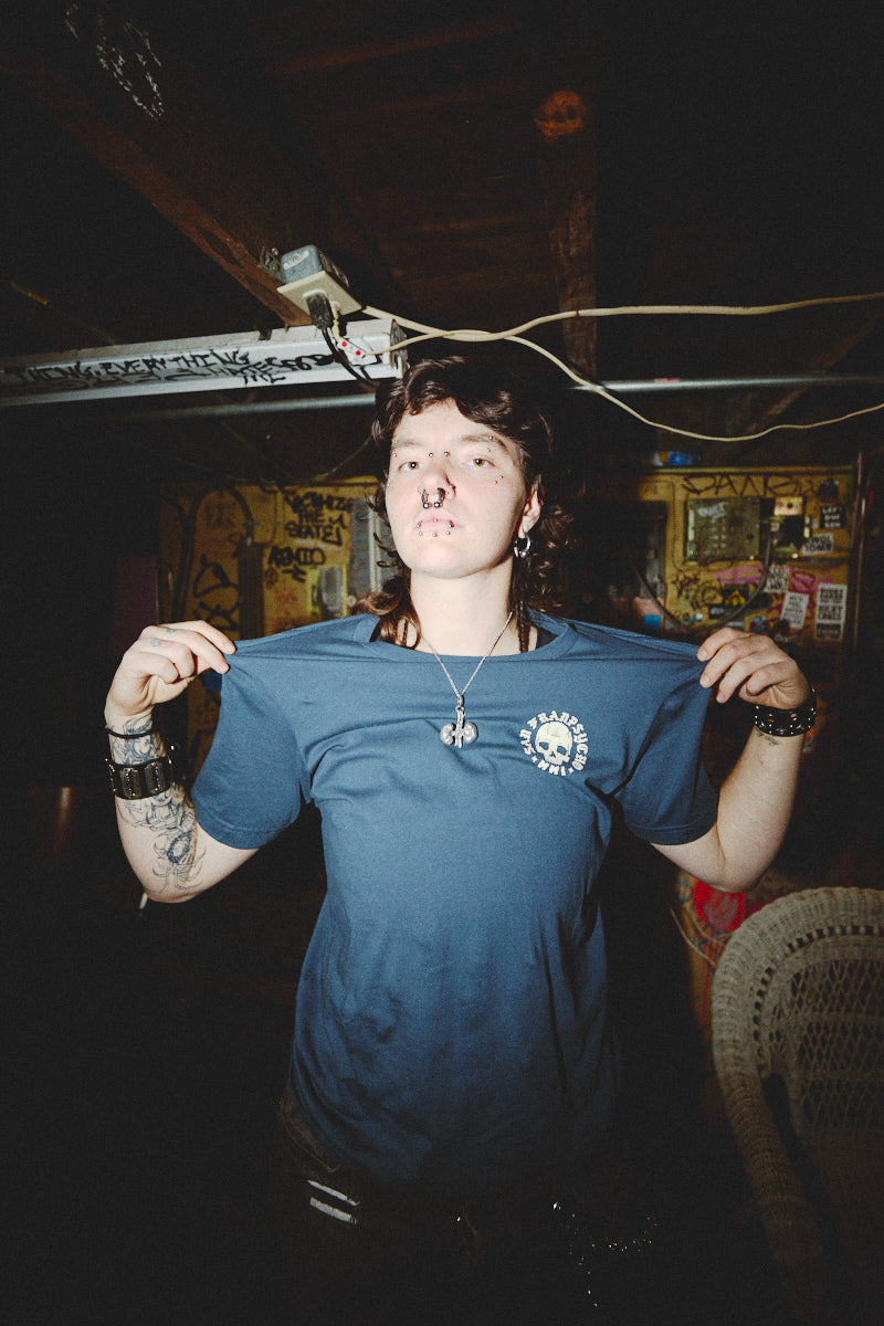 Amidst a graffiti-covered room, a person adorned with piercings and tattoos holds up a hand-printed BBNW Skull Tee in electric blue, effortlessly capturing the rebellious essence of Punk Rock 2023.