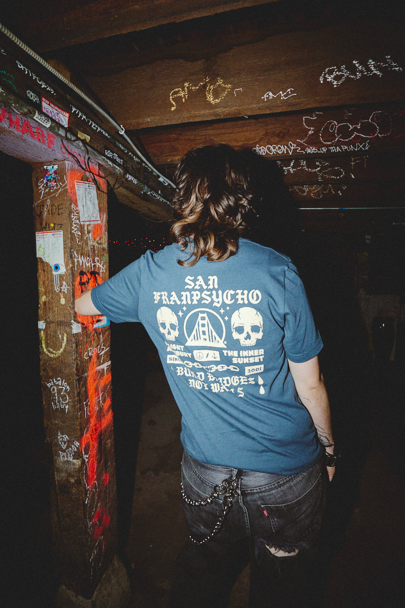 A person wearing a hand-printed BBNW Skull Tee leans against a graffiti-covered beam in the dimly lit space, embodying the gritty essence of Punk Rock 2023.