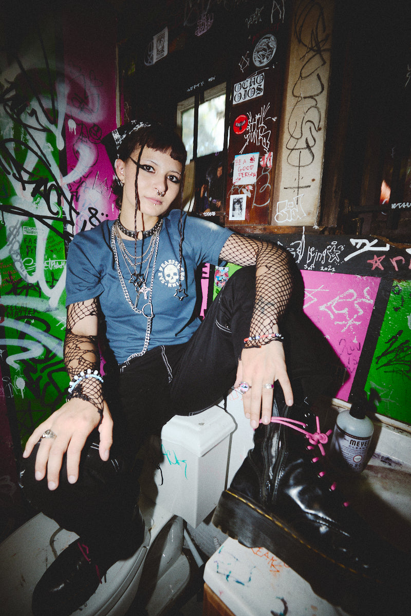 A person in punk attire relaxes in a graffiti-covered room, donning a hand-printed BBNW Skull Tee with fishnet sleeves, a nod to Punk Rock 2023. Black boots complete the edgy ensemble under the glow of rebellion.
