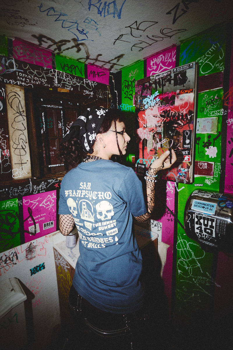 In a graffiti-covered room, a person in a BBNW Skull Tee adjusts a poster on the wall.