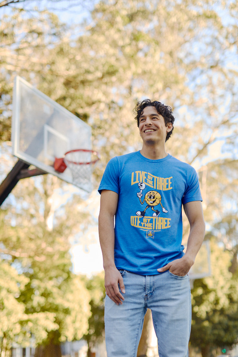 A smiling man in a "Live by the Three Blue Tee" stands near a basketball hoop outdoors, exuding the confidence of a true Warriors fan.