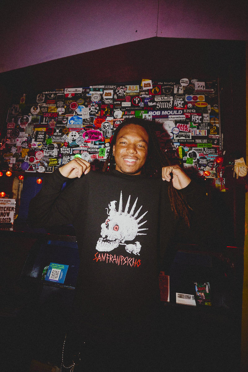 In a room adorned with stickers, a person beams at the camera while clutching the Rat Skull Crewneck from the Sanfranpsychco Fall 2023 Collection, perfectly embodying punk rock style.
