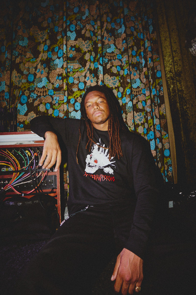 A person with long hair sits in front of floral curtains, dressed in dark pants and a black sweatshirt featuring the Rat Skull Crewneck design from the Fall 2023 Collection. This edgy piece adds a touch of punk rock flair to their style.