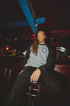 A person with long hair sits on a bar stool, wearing a gray and black BBNW Skull Patched Raglan Crewneck, in a dimly lit room with red lights.