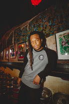 A person with dreadlocks casually leans on a keg in a dimly lit, decorated room filled with framed art on the walls, exuding a laid-back punk rock vibe while wearing the BBNW Skull Patched Raglan Crewneck.