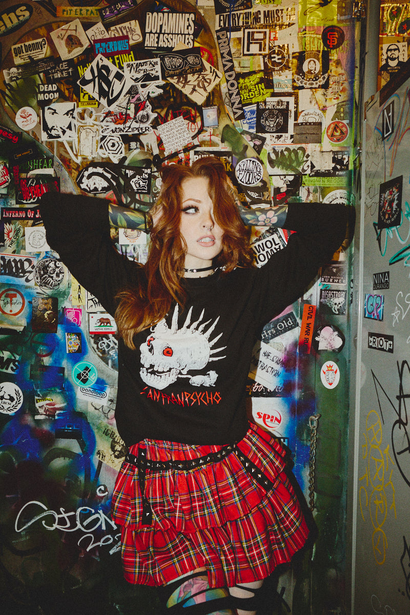 A red-haired person wearing a plaid skirt leans against a graffiti-adorned wall, showcasing the Rat Skull Crewneck. Embodying the bold essence of the Fall 2023 Collection, their outfit exudes Punk Rock rebellion.