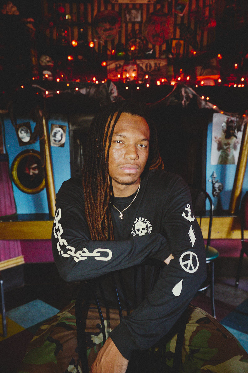 A man with long dreadlocks sits on a chair in a colorful room adorned with string lights and eclectic wall decor, wearing the BBNW Skull Long Sleeve shirt.