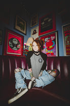A person with tattoos sits cross-legged on a red couch, wearing a BBNW Skull Patched Raglan Crewneck. The room is adorned with colorful posters, echoing a punk rock vibe.