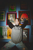A person in a BBNW Skull Patched Raglan Crewneck strikes a playful expression in front of framed posters on a blue wall.