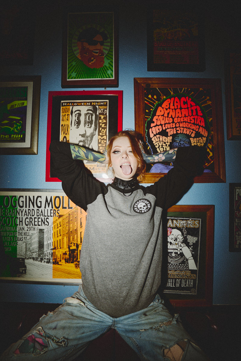 A person in a BBNW Skull Patched Raglan Crewneck strikes a playful expression in front of framed posters on a blue wall.