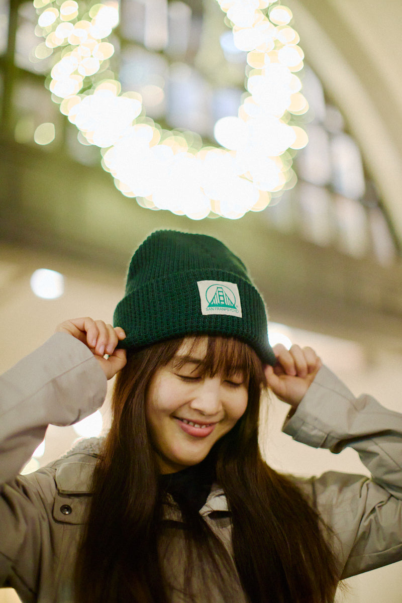 A smiling woman wears the Forest Green Waffle Beanie w/ Natural/Green Logo while standing indoors under a glowing circular holiday light display.