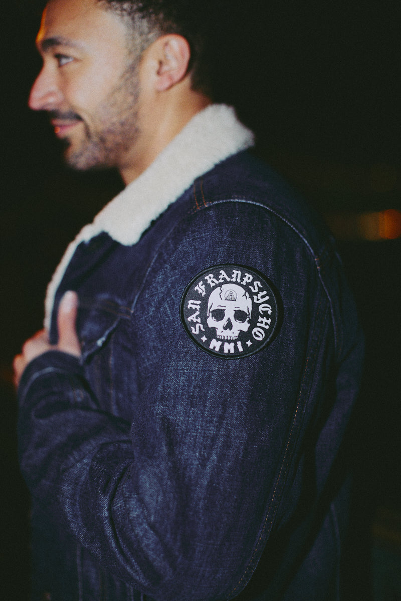 A man stands against a dark background wearing a denim jacket featuring the Punk Skull Patch embroidered on the sleeve.