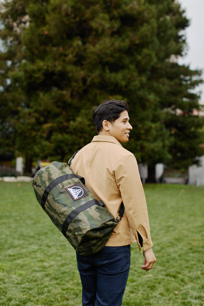 A person carrying an SFP Patched Duffle Bag, made of durable polyester and featuring a removable shoulder strap, walks on a grassy field with trees in the background.