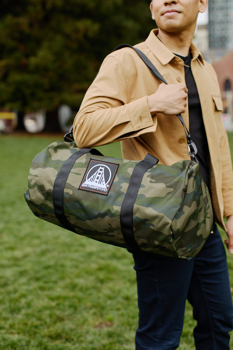 A person in a beige jacket is standing on grass with the SFP Patched Duffle Bag, featuring a camo design and made of durable polyester, slung over their shoulder.