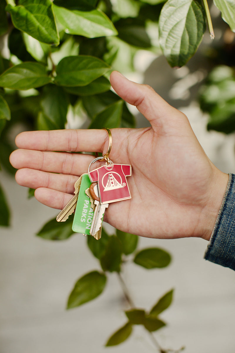 A hand holding a set of keys attached to the Logo Tee Keychain, designed in the shape of a red T-shirt, amidst vibrant green foliage.