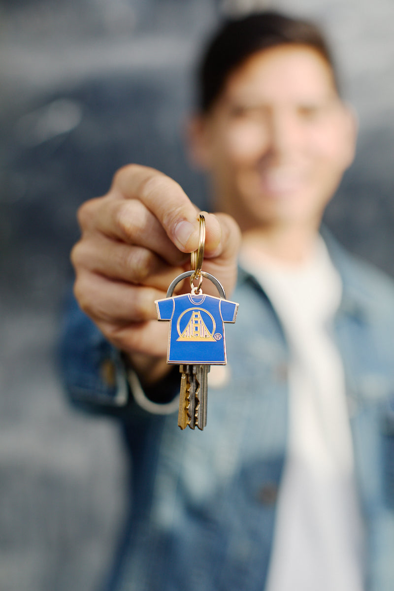 A person is holding keys adorned with a vibrant Logo Tee Keychain shaped like a shirt, smiling in the background.