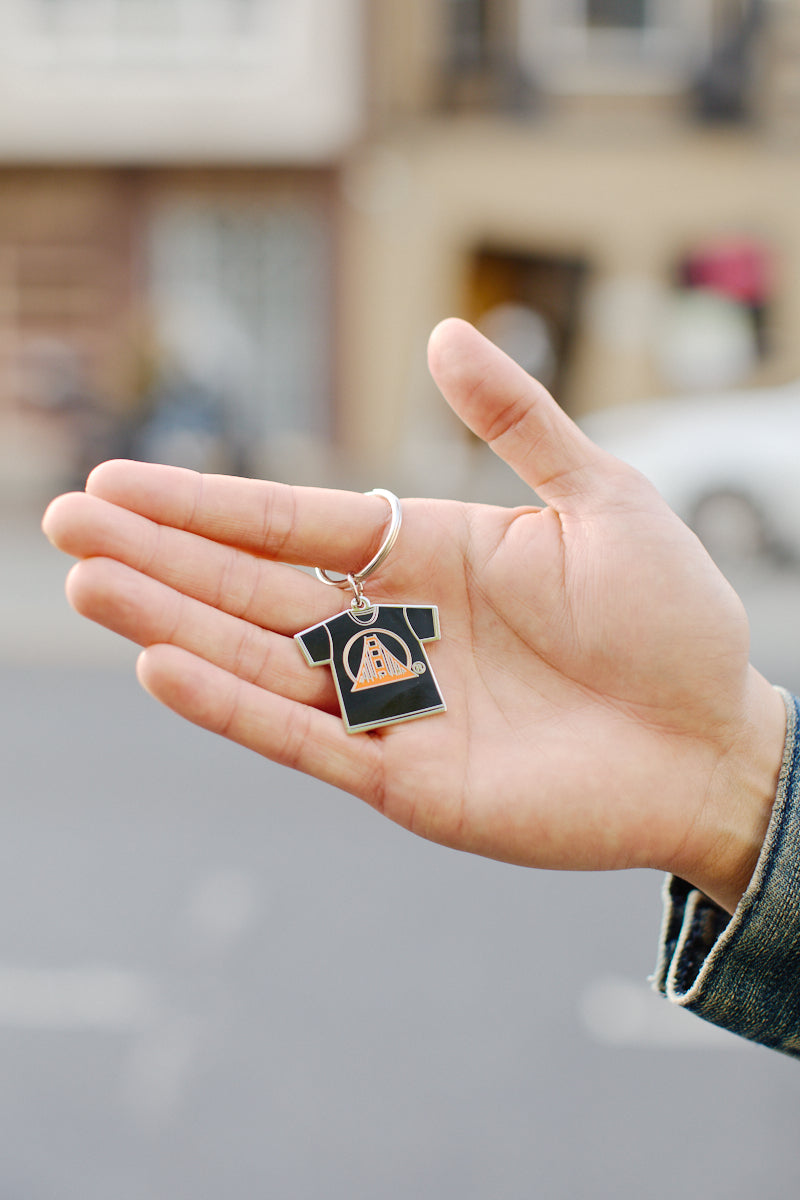 A hand grasps the Logo Tee Keychain, meticulously shaped like a black t-shirt. The design, enhanced by enamel filling at its center, adds a touch of flair to this charming accessory.