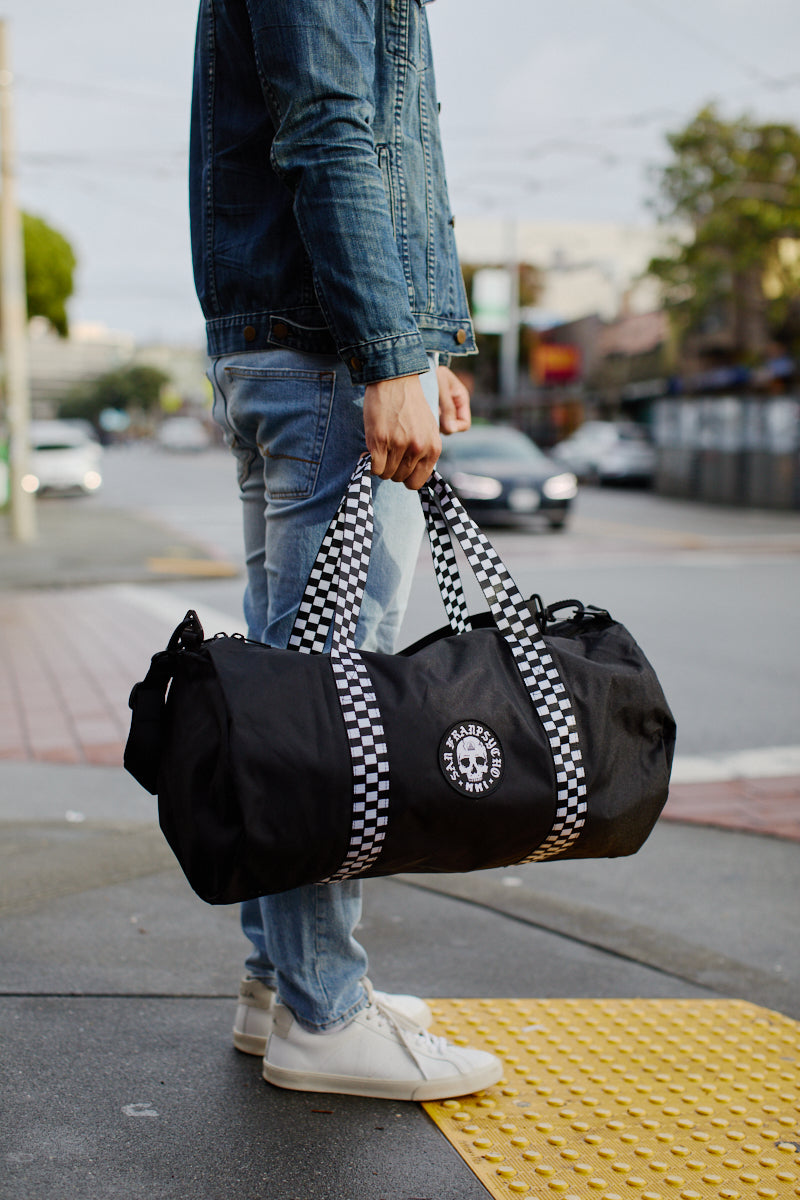 A person is standing on a street corner, holding the Skull Patched Duffle Bag, which features checkered straps and a removable shoulder strap, crafted from durable polyester.
