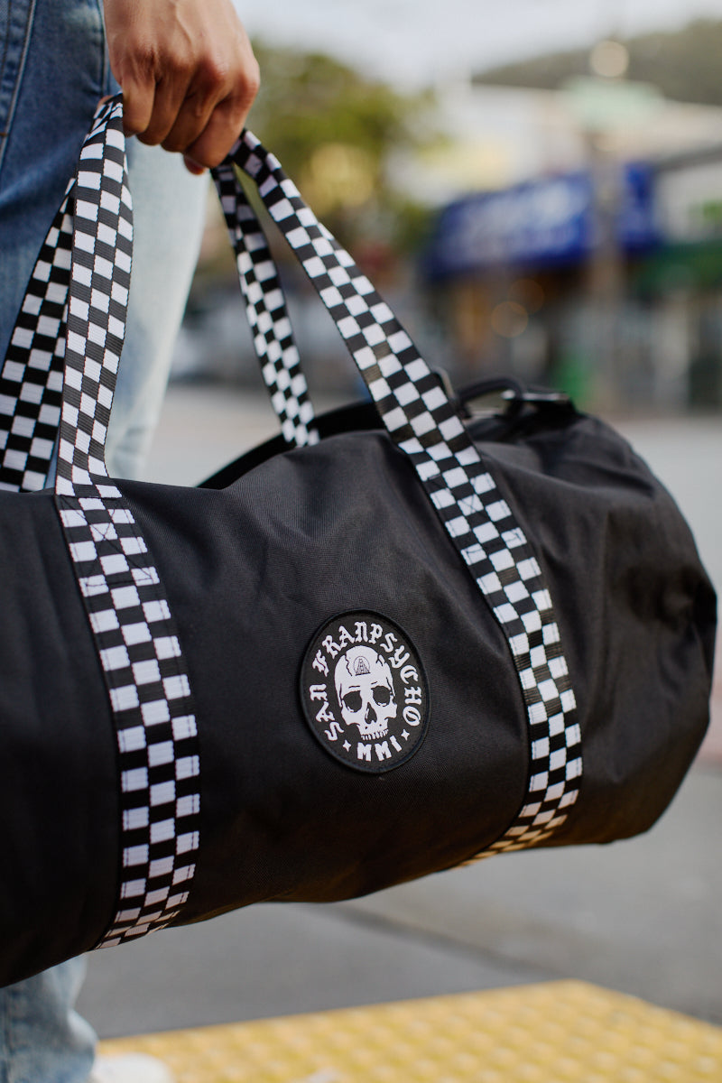 A person holds the Skull Patched Duffle Bag, a black bag made of durable polyester with checkered straps and a striking skull logo.