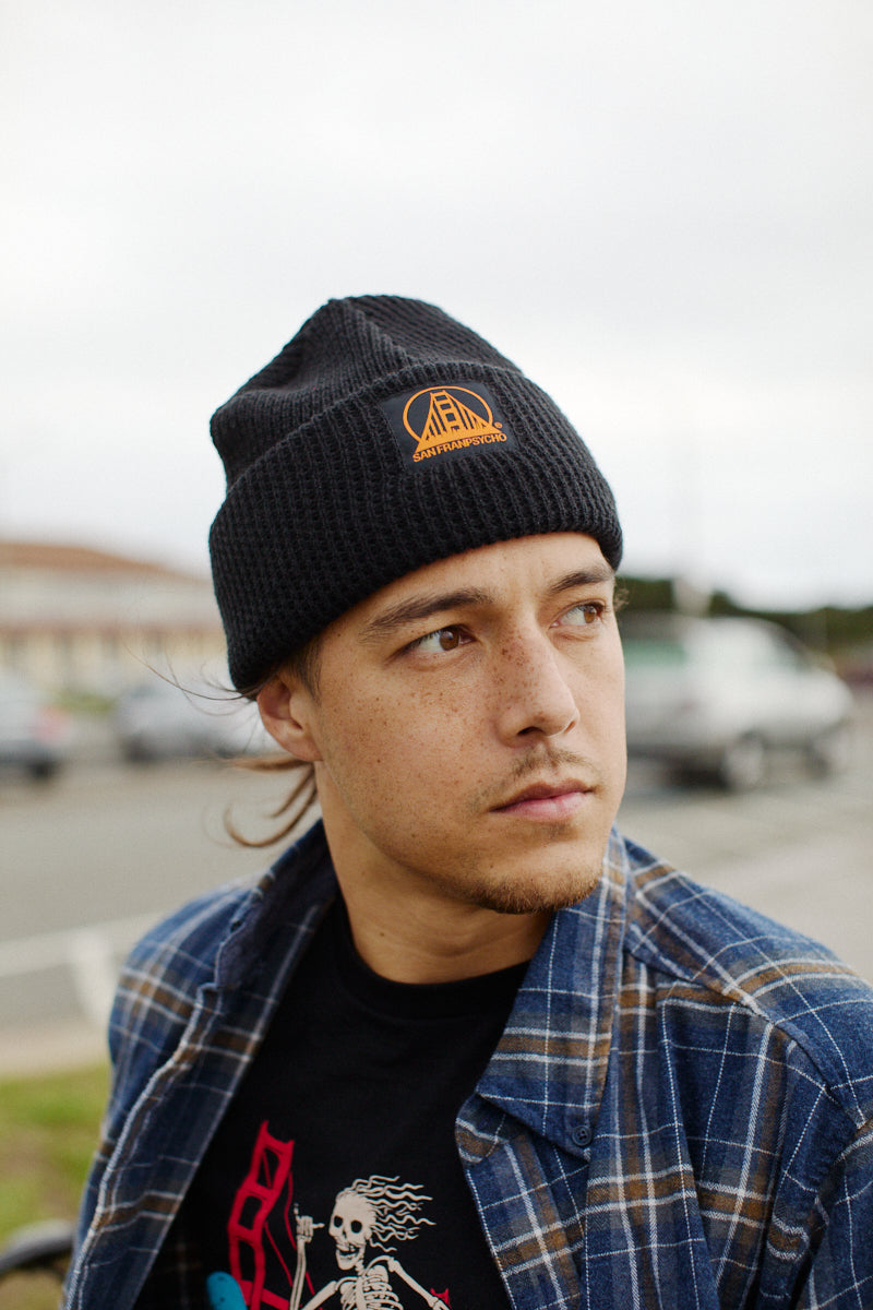 A person in a plaid shirt and a Black Waffle Beanie with the Black/Orange SFP Logo is looking sideways outdoors.